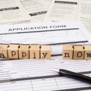 Application forms and a pen laid out on a desk with letter stamps forming the words 'apply now'
