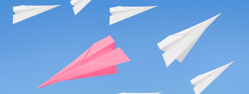 White paper airplanes flying to the right while one pink paper airplane is flying to the left against a blue background