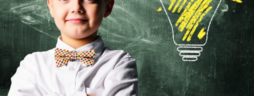 Kid in a dress shirt and bowtie standing in front of a chalkboard with a lit up lightbulb drawn on it